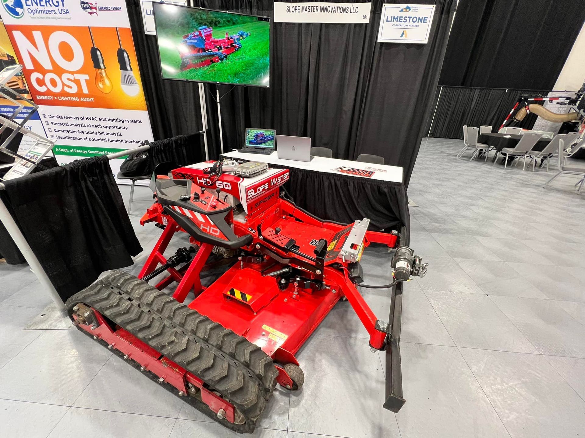 Red tracked lawn mower on display at a trade show. A screen shows the mower in use.