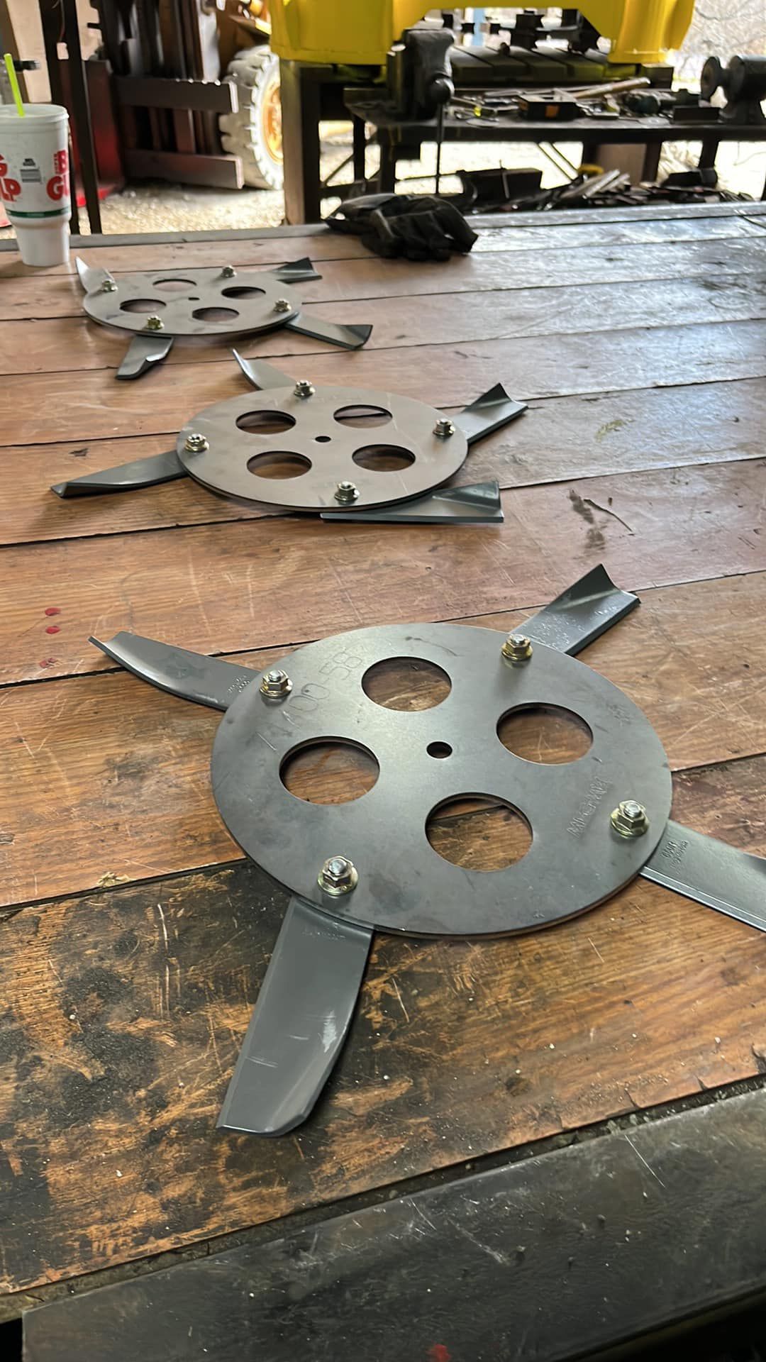Three metal mower blades with circular hubs and curved cutting blades on a wooden surface.