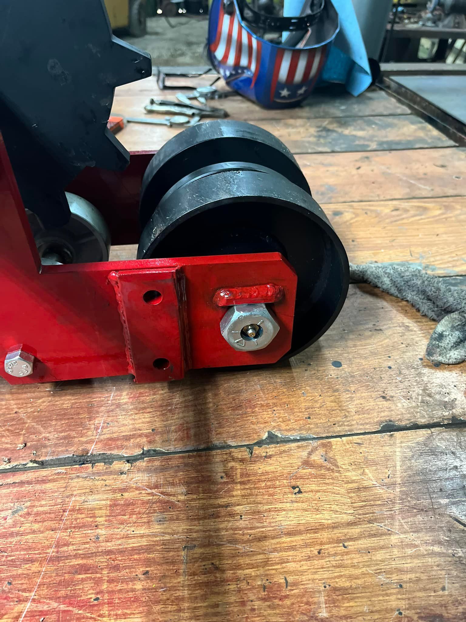 Red and black metal machine with wheels on a wooden surface, with a weld helmet in background.