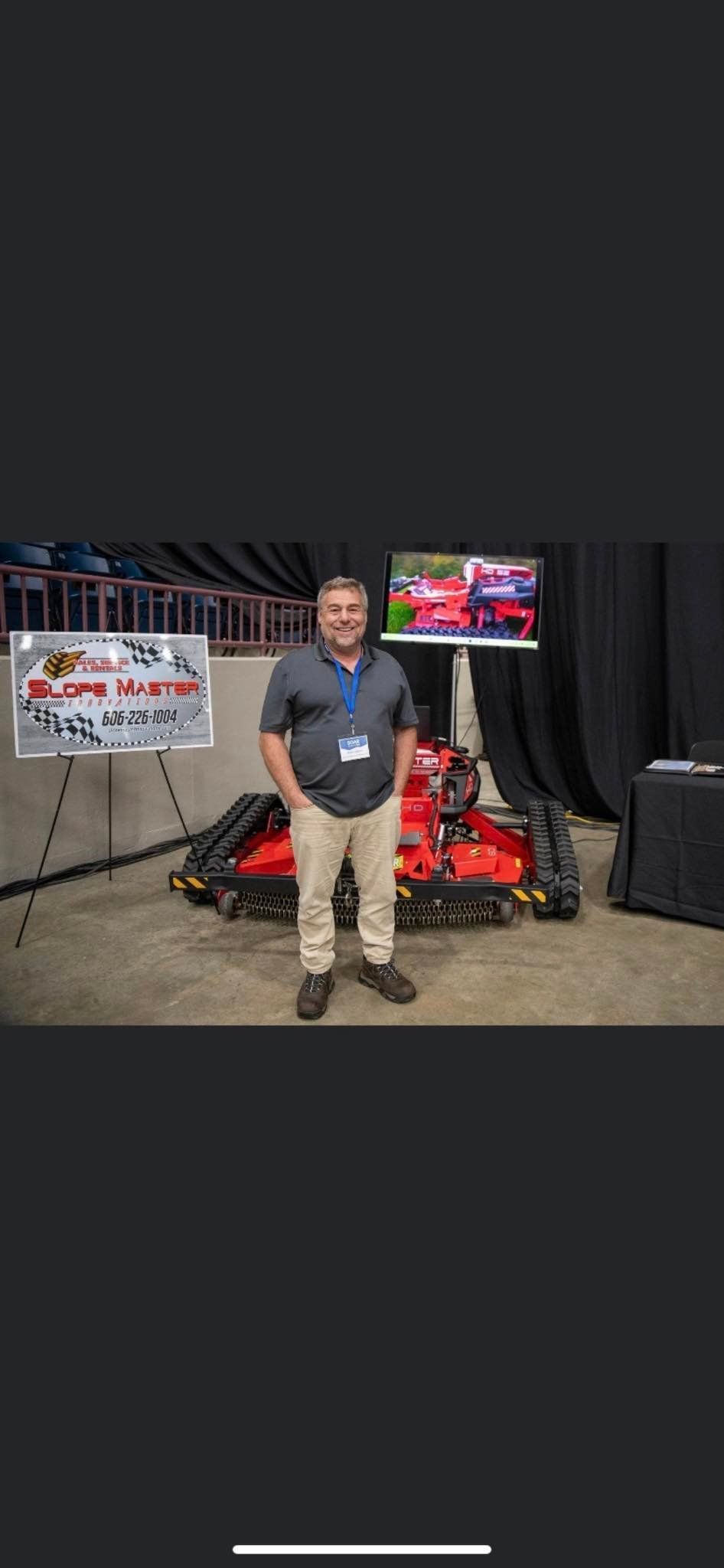 Man in tan pants stands in front of a red robot, with a sign to the left and a screen in the back.