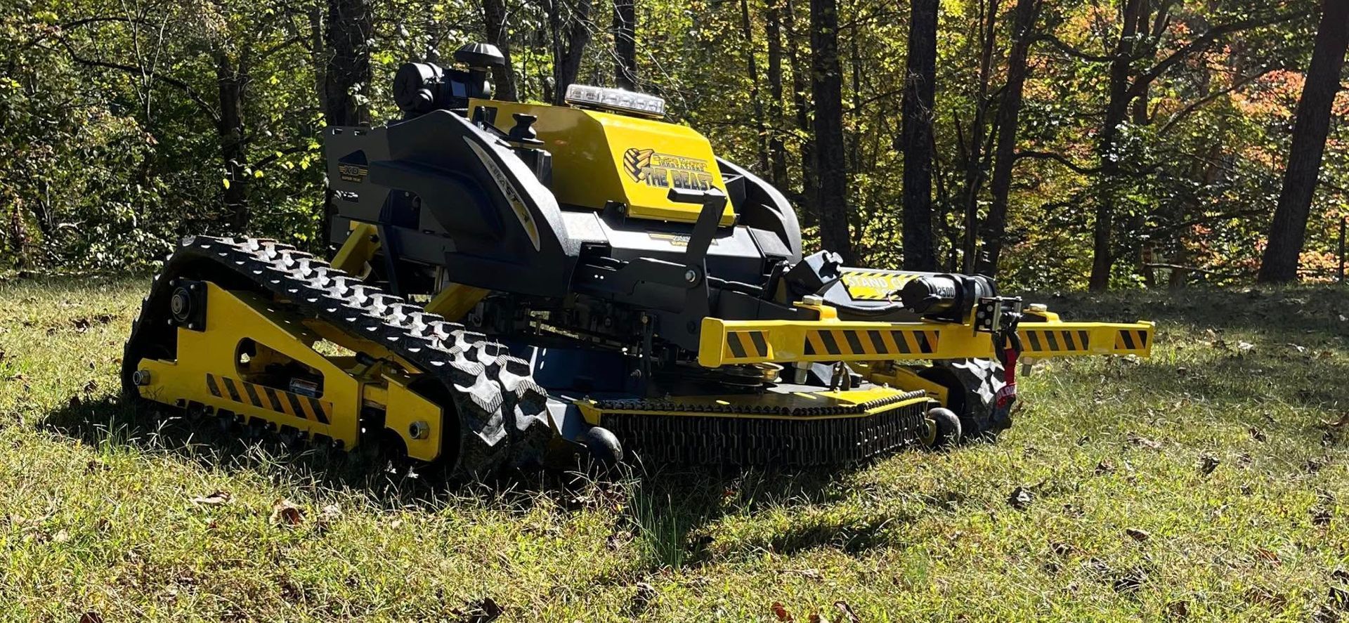 A yellow and black tracked mower in a grassy area, with trees in the background.