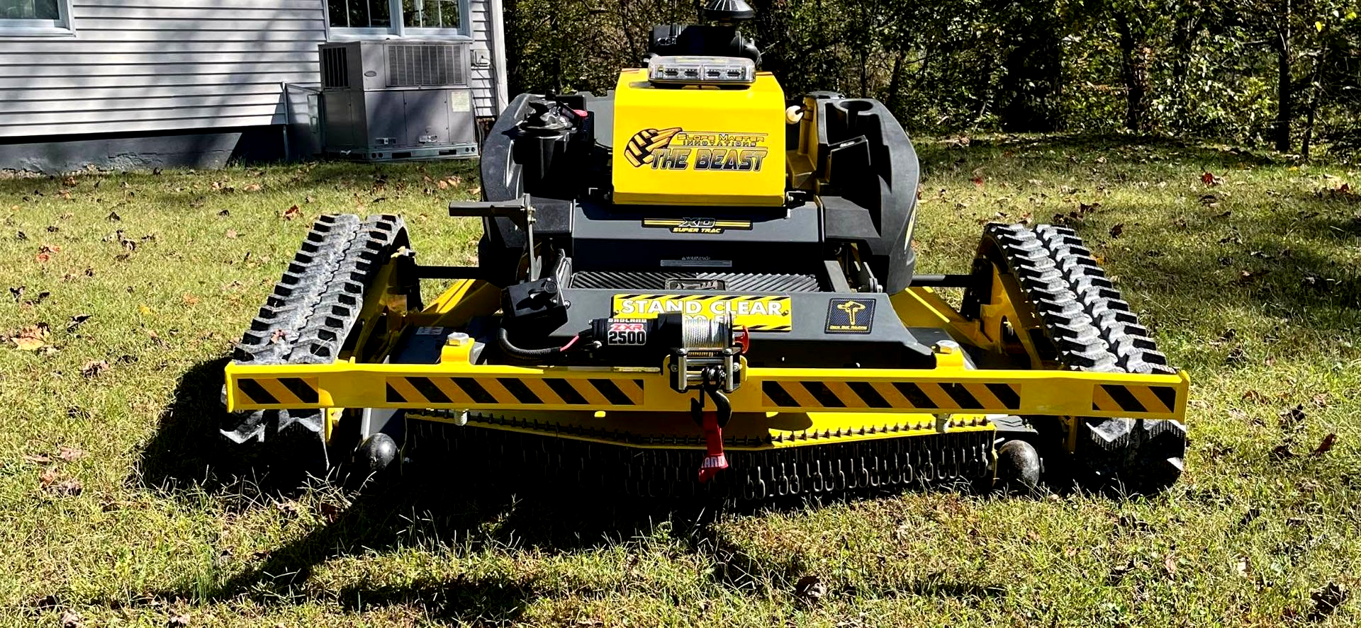 A yellow, remote-controlled track-driven brush mower parked on a residential lawn.
