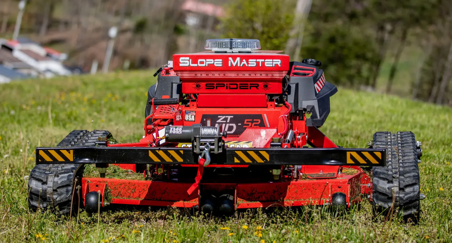 Red robotic lawnmower, Slopemaster Spider, on a grassy slope.  It has treads and flashing light on top. 