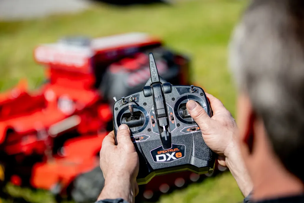A person holds a black remote control, operating a red, tracked vehicle outdoors on a grassy field.