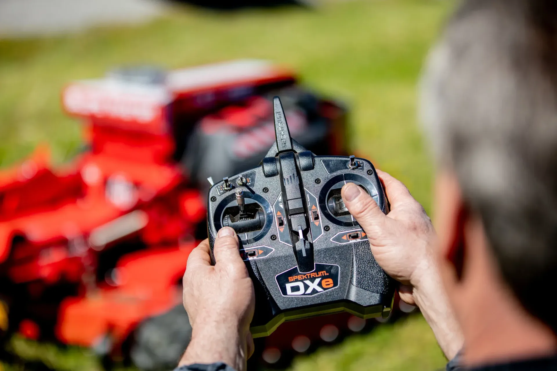Person holding a black remote control, operating a red RC vehicle outdoors on grass.