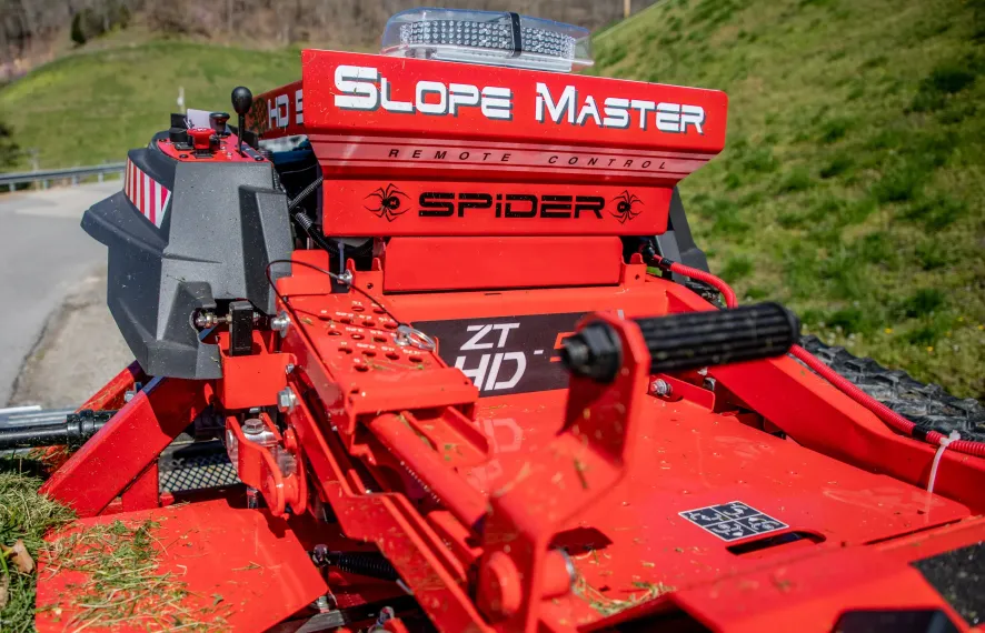 Red Spider Slope Master remote-controlled mower on a grassy slope, beside a road.