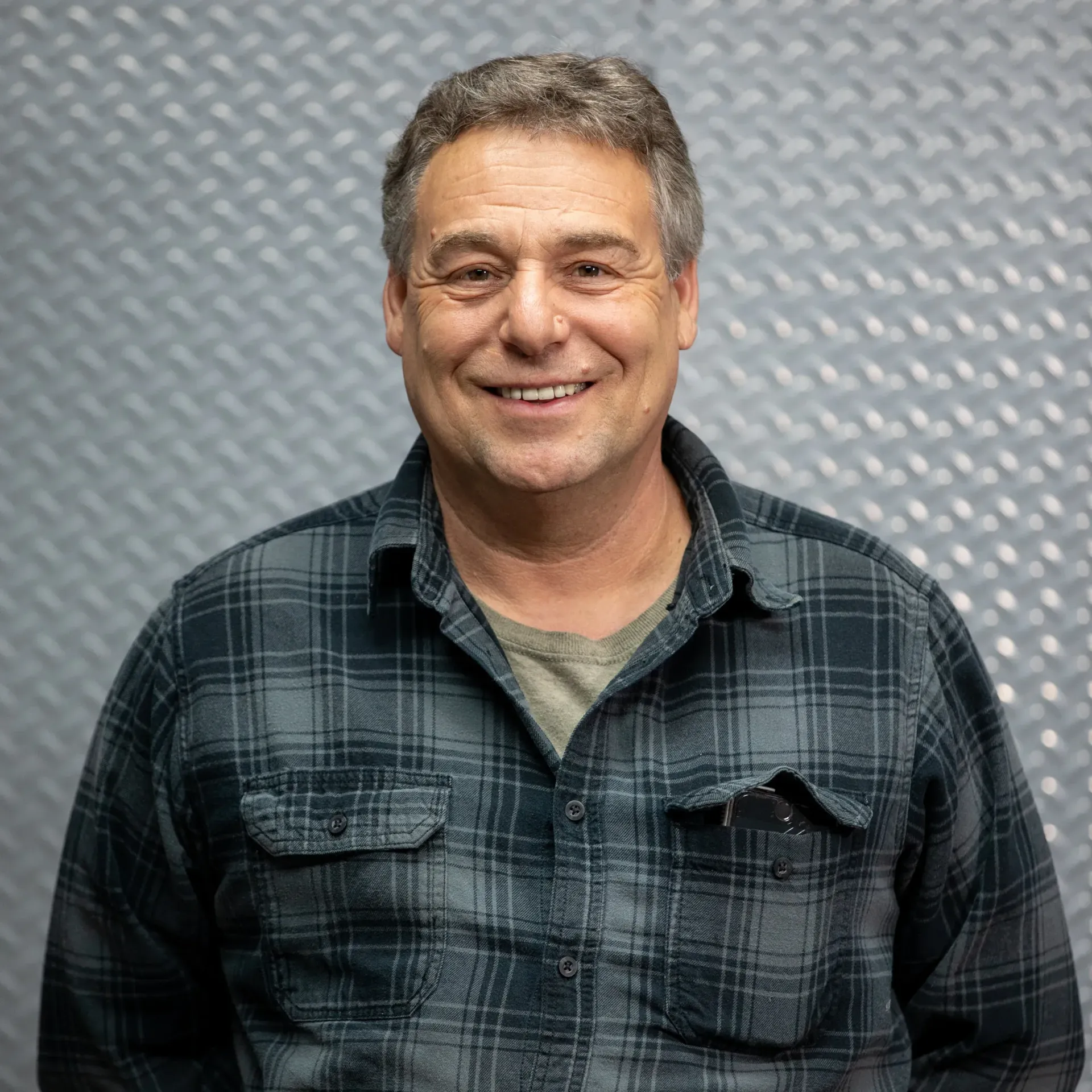 Middle-aged man with graying hair smiles, wearing a plaid shirt in front of a metallic wall.