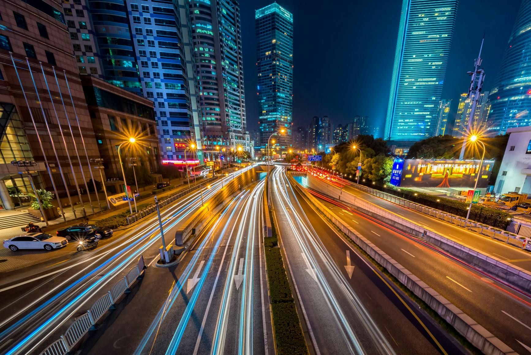 Night view of a busy highway with long exposure light trails, flanked by tall, illuminated skyscrapers in a city.
