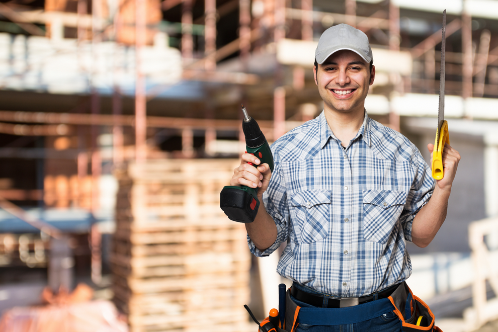 Smiling construction worker holding a drill and saw on a construction site.