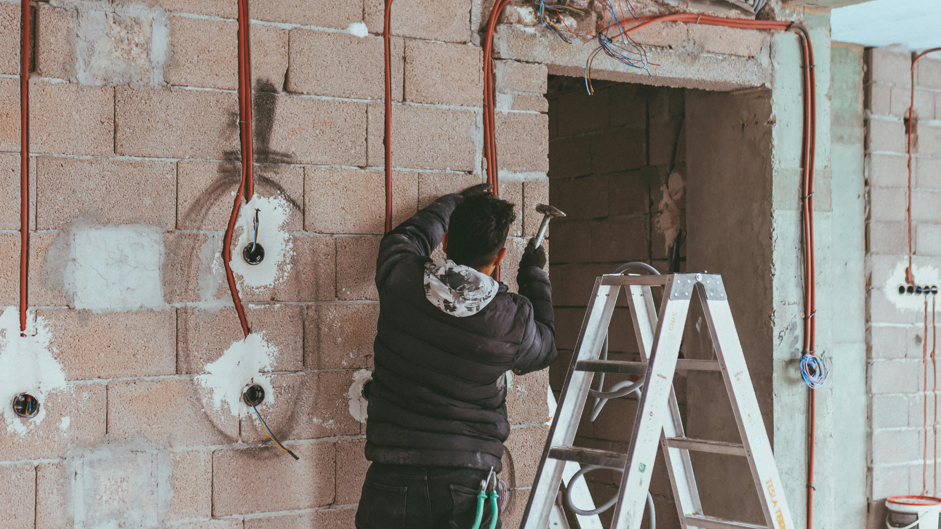 Person on ladder installing electrical wiring in a cinder block wall during construction.