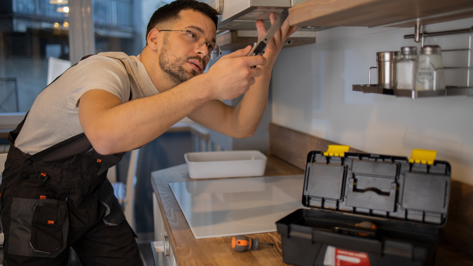 Man in overalls repairs a kitchen fixture with a screwdriver, toolbox nearby.