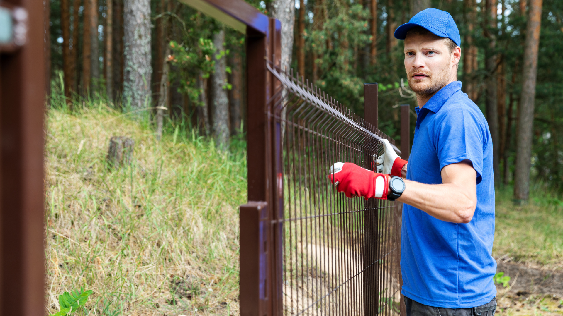 Man in blue shirt and cap installing a brown fence outdoors, wearing red gloves.