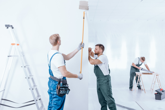 Three painters working on a white room: one painting the ceiling, one holding a board, and one sawing wood.