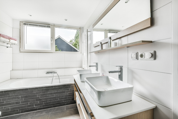 White bathroom with double sinks, tub, and window. Light wood vanity and accent tile.