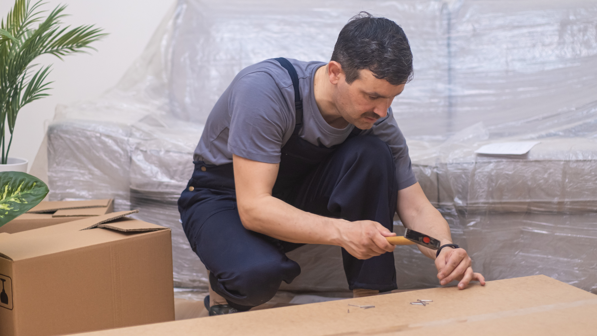 Man kneeling, hammering cardboard. Sofa covered in plastic, boxes nearby. Indoor setting.