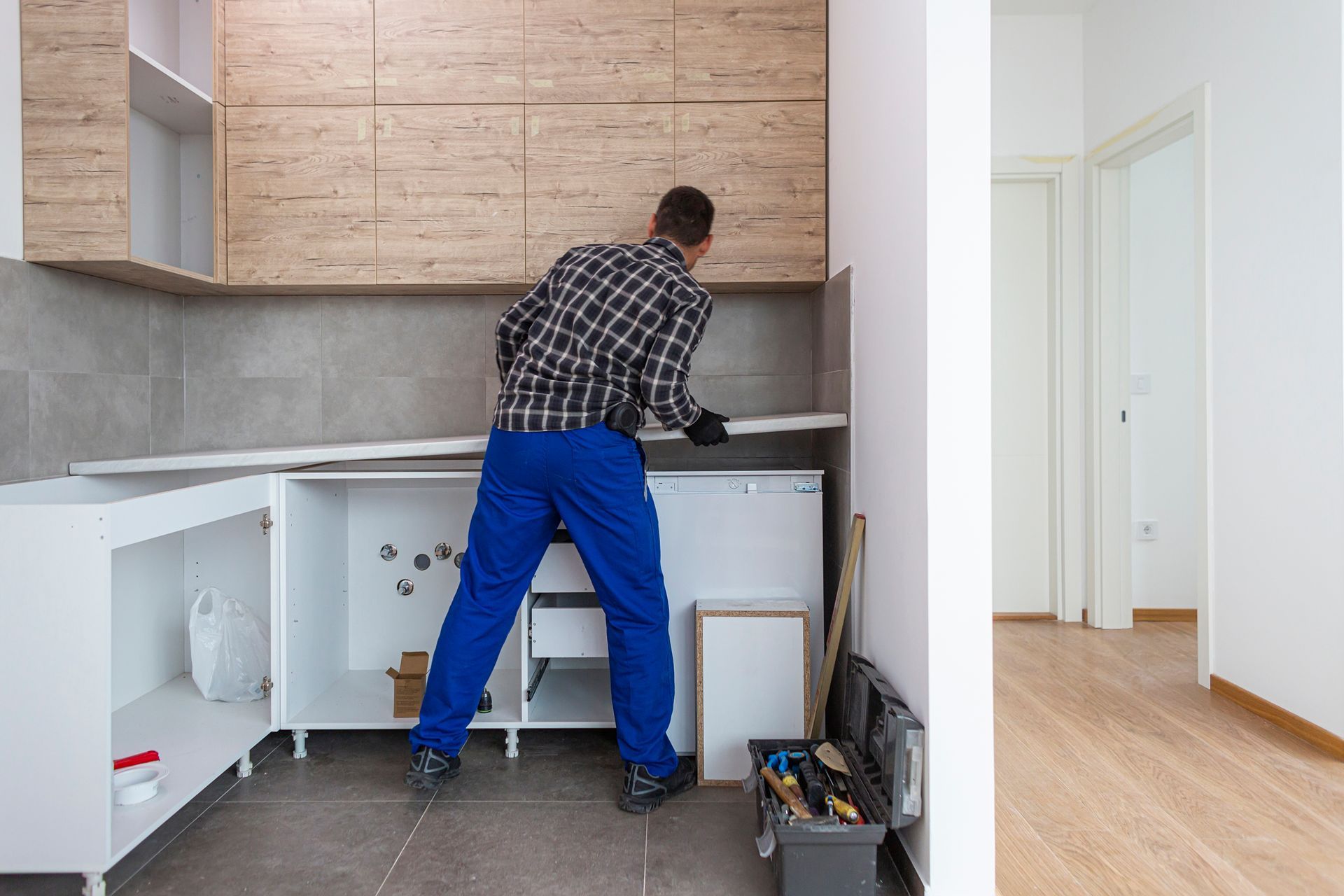 Man in blue overalls installing a kitchen cabinet, inside a room.