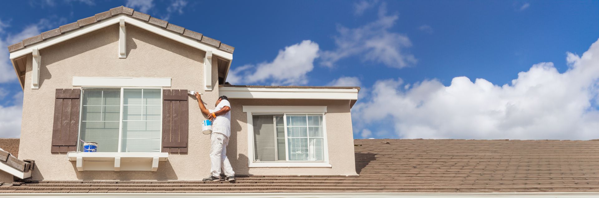 A house painter is working on a home's exterior under a bright blue sky.