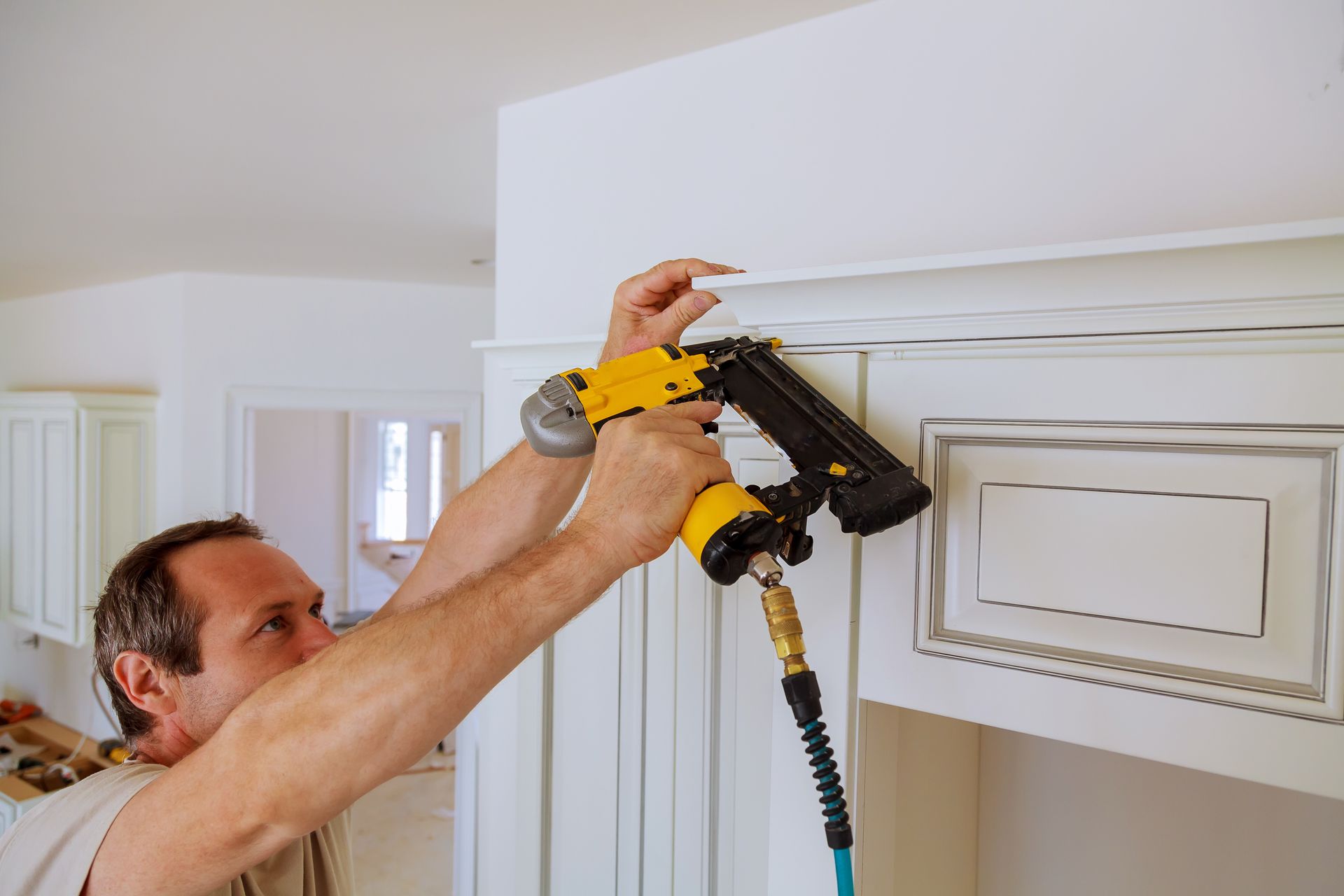 Man using a yellow and black nail gun to attach white trim to a white cabinet.