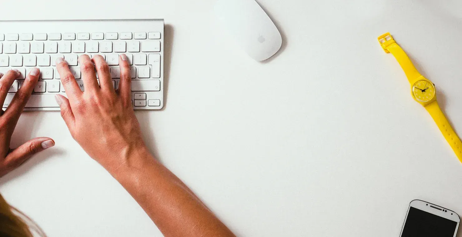 A person is typing on a white keyboard on a white desk.