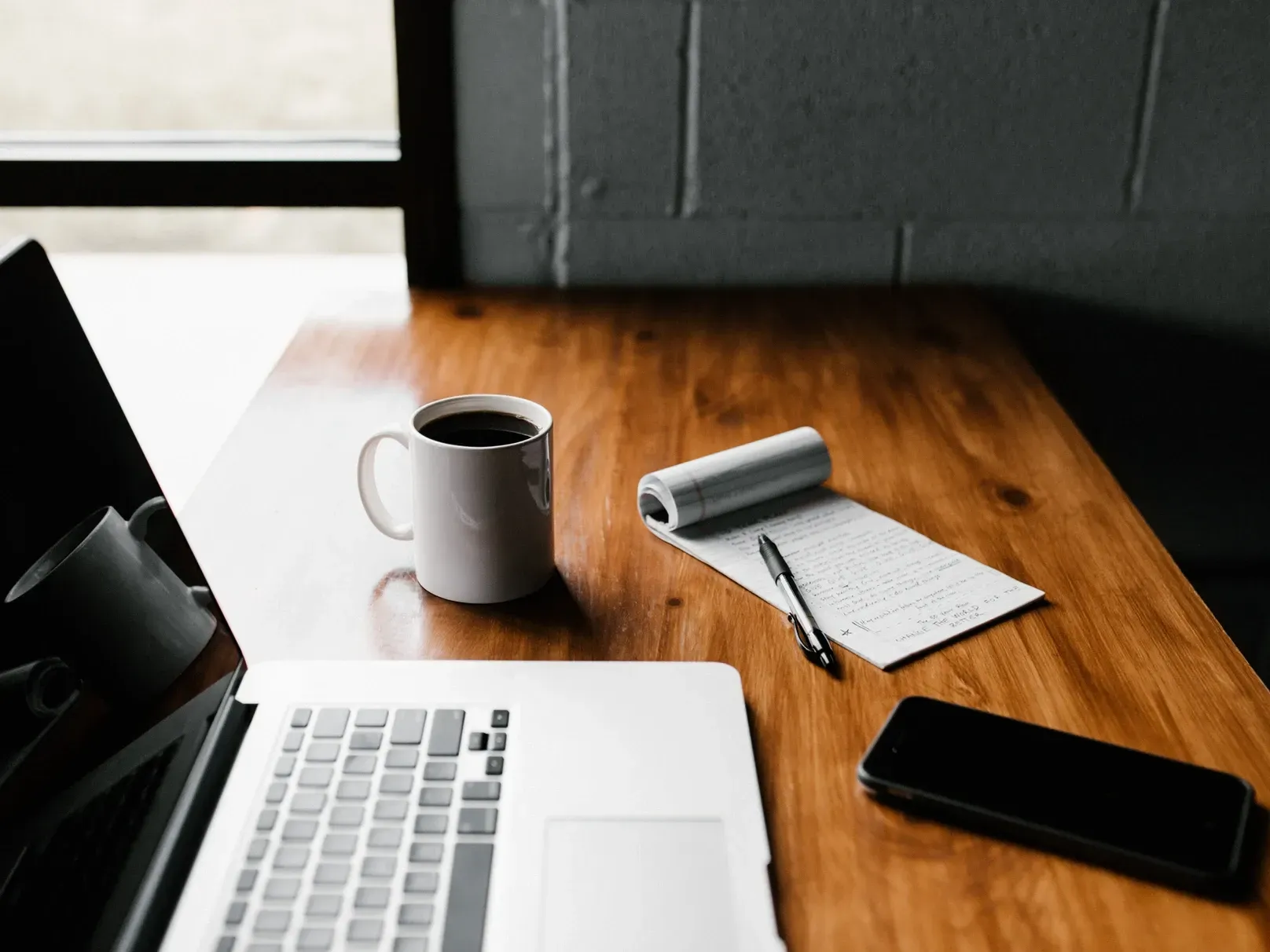 A wooden desk with a laptop a cup of coffee a notebook and a cell phone.