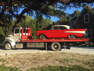 Red classic car on a flatbed tow truck. White roof, red body. Outdoors under a tree, beside a house.