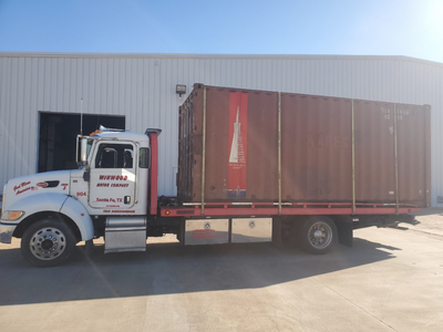White semi-truck carrying a large brown shipping container in front of a white building.