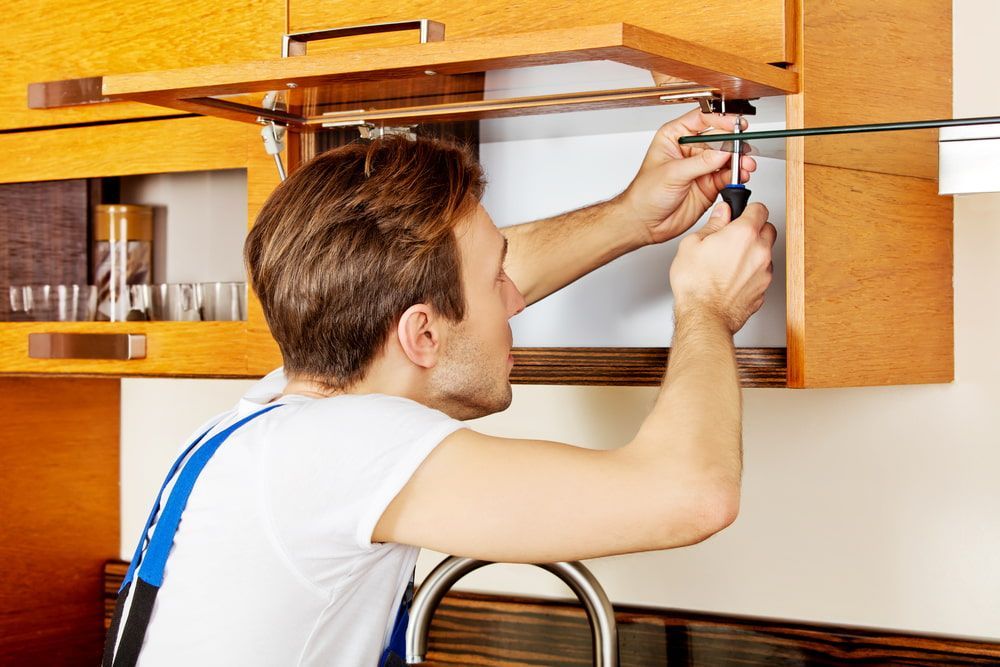 A Man Is Fixing A Cabinet In A Kitchen With A Screwdriver — CBY Carpentry & Renovations In Dubbo, NSW