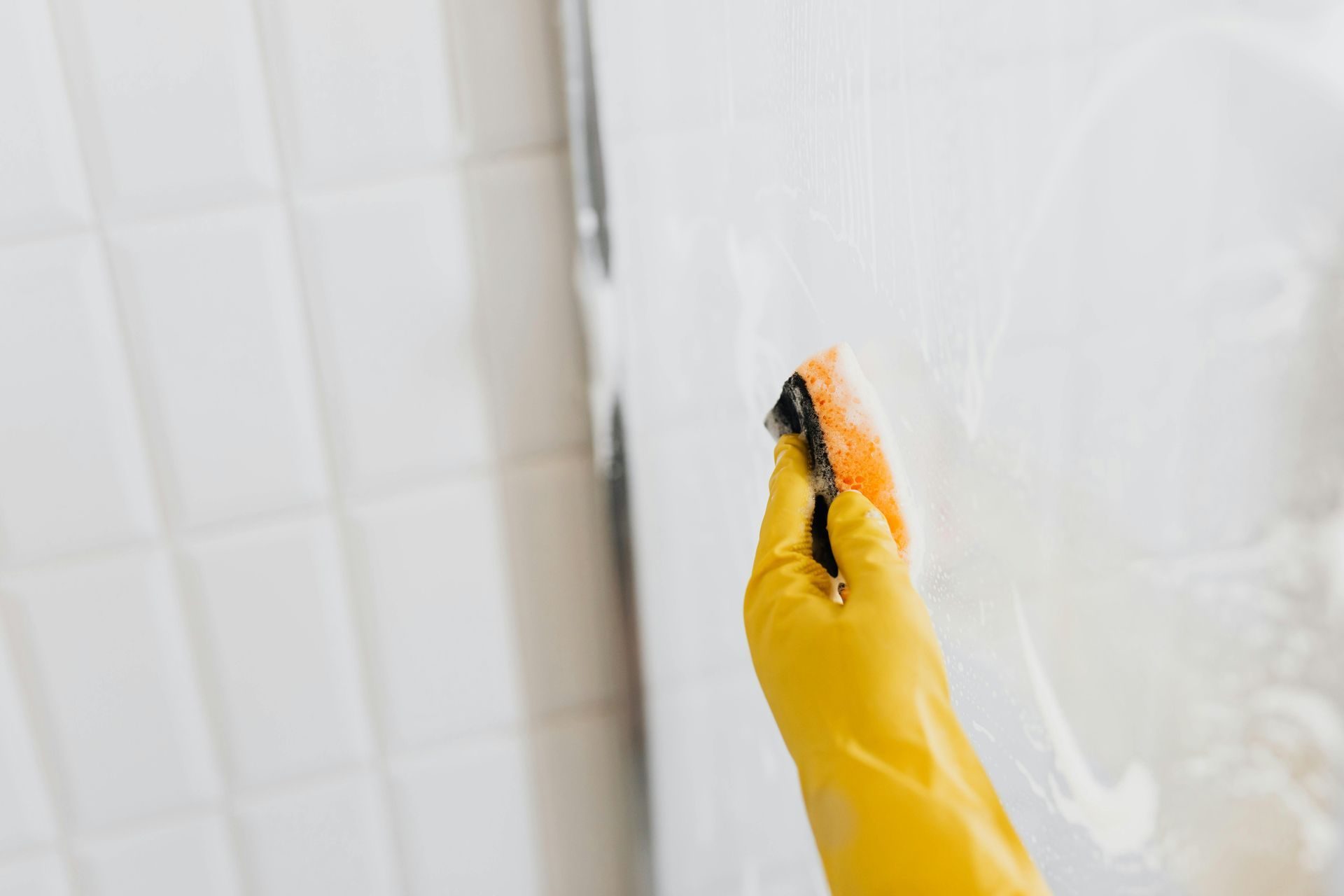 Hand in yellow glove scrubbing a white surface with a sponge. White tile wall on the left.