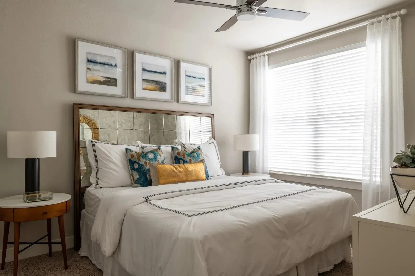 A stylish bedroom featuring a bed with decorative pillows, a lamp, and a window with sheer curtains.