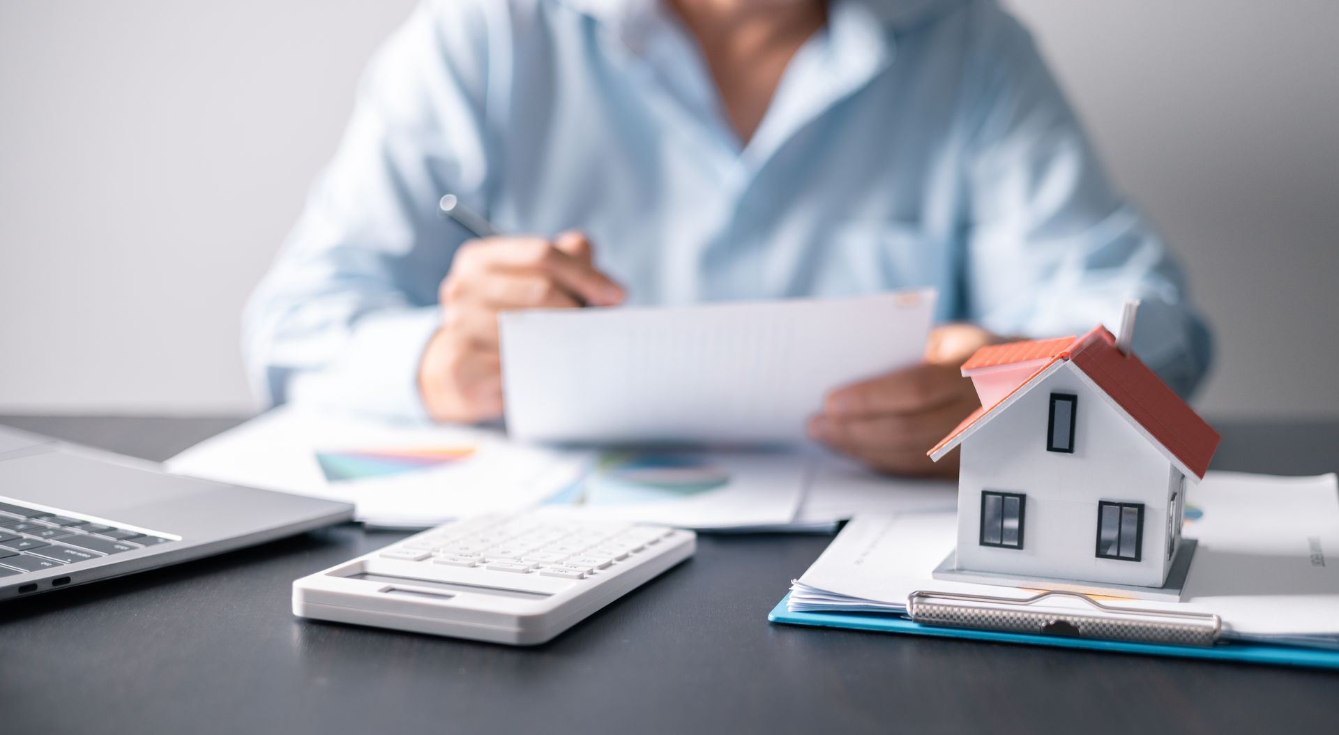 Person reviewing documents with a calculator, laptop, and model house on a desk.
