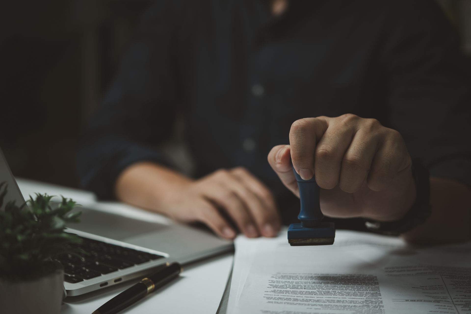 Person stamps a document with a blue stamp on a desk with a laptop and pen.