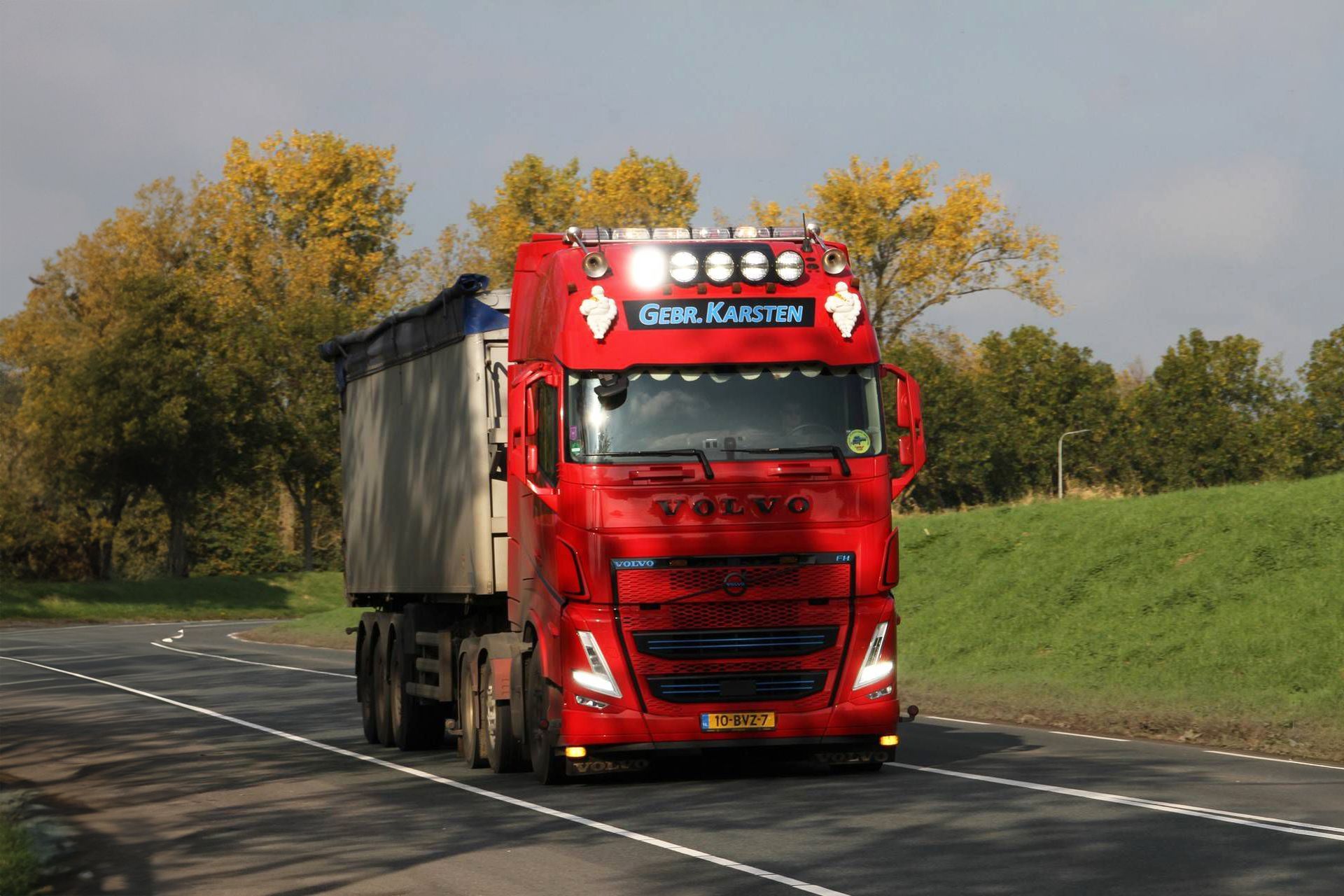 Rode Volvo-vrachtwagen met oplegger rijdt over de weg. Bomen met gele bladeren op de achtergrond.