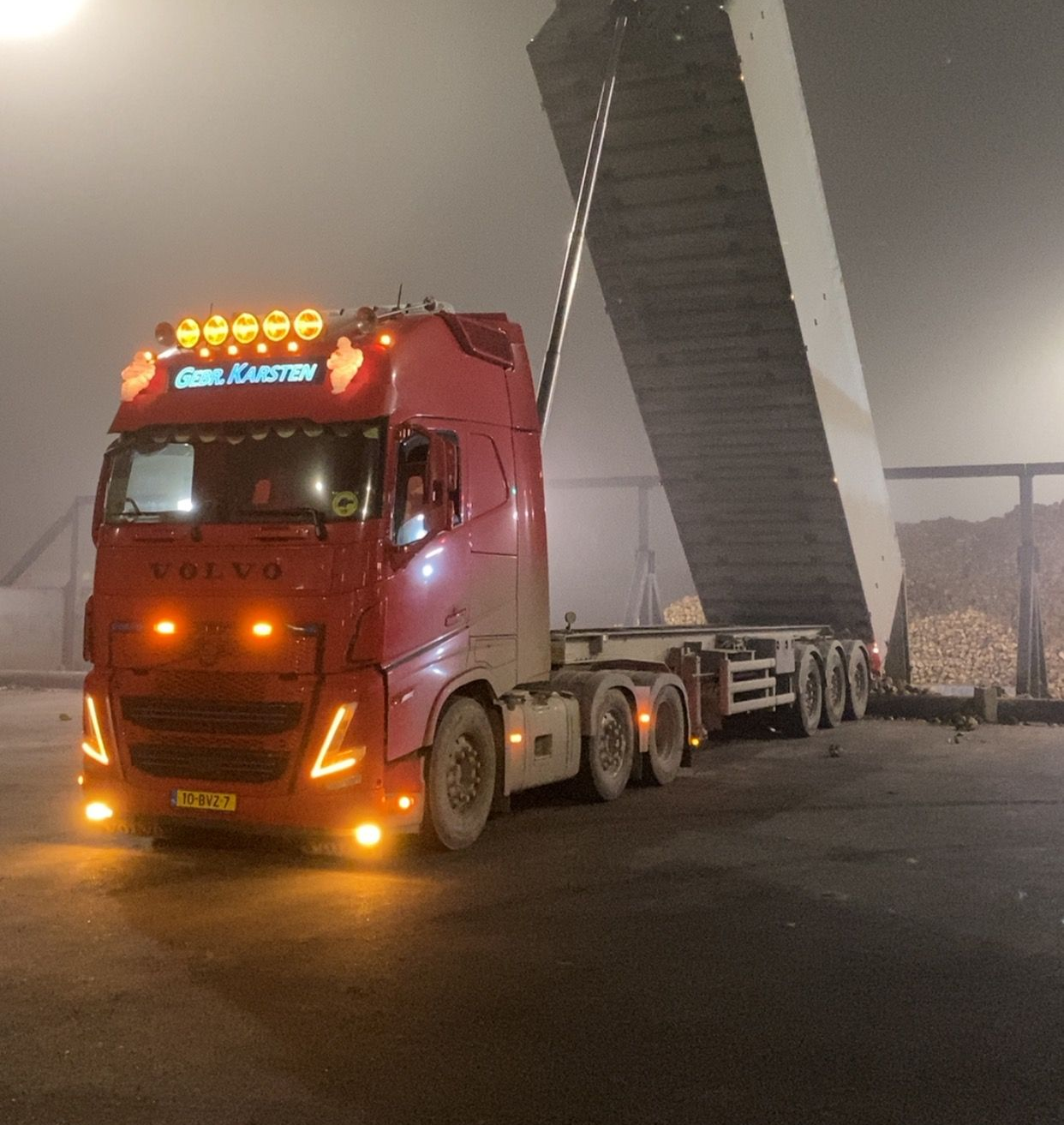 Rode Volvo-vrachtwagen met verhoogde oplegger die een lange betonnen constructie vervoert in de nacht.