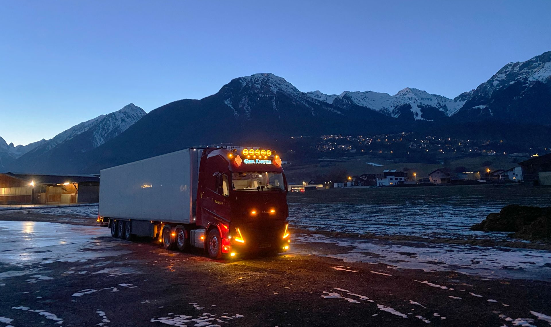 gebr-karsten-transport rode vrachtwagen met verlichting in besneeuwd berggebied bij stad in schemering