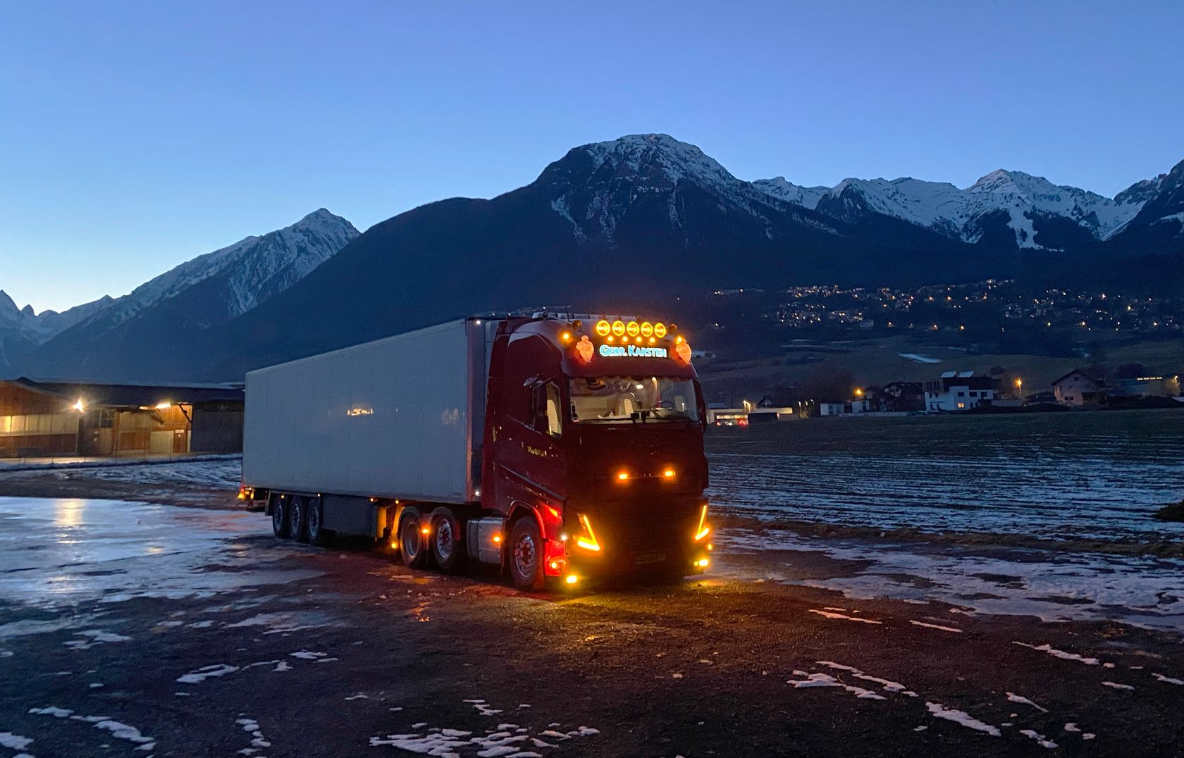 Rode vrachtwagen met aanhanger op een besneeuwd terrein in de schemering, met bergen op de achtergrond.