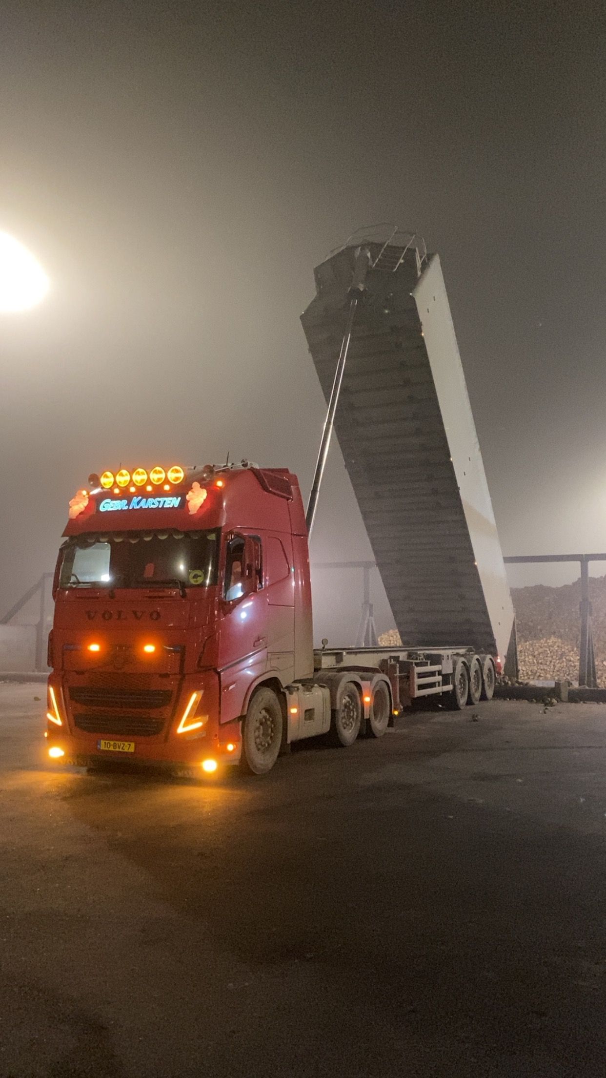 Een rode vrachtwagen staat gekanteld en laadt een hoge stapel grijze materialen uit in een mistige, donkere omgeving.