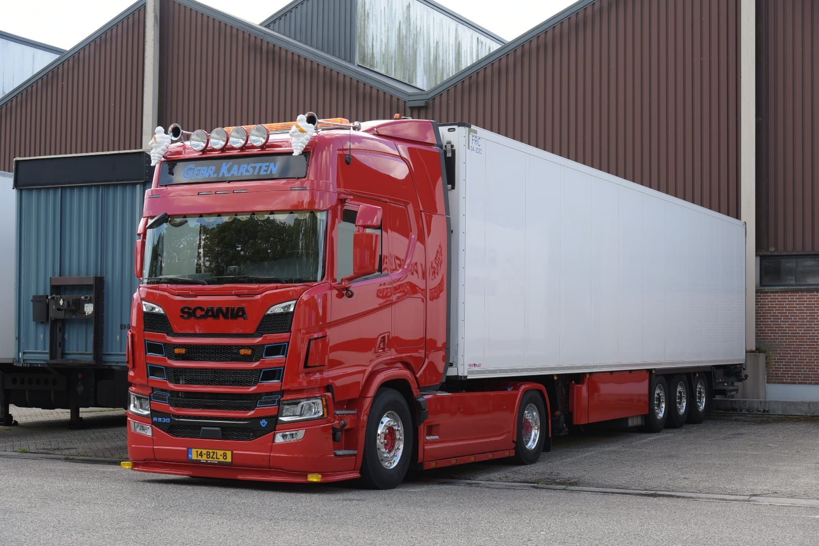 Rode Scania vrachtwagen met witte oplegger geparkeerd voor bruine en blauwe industriële gebouwen.