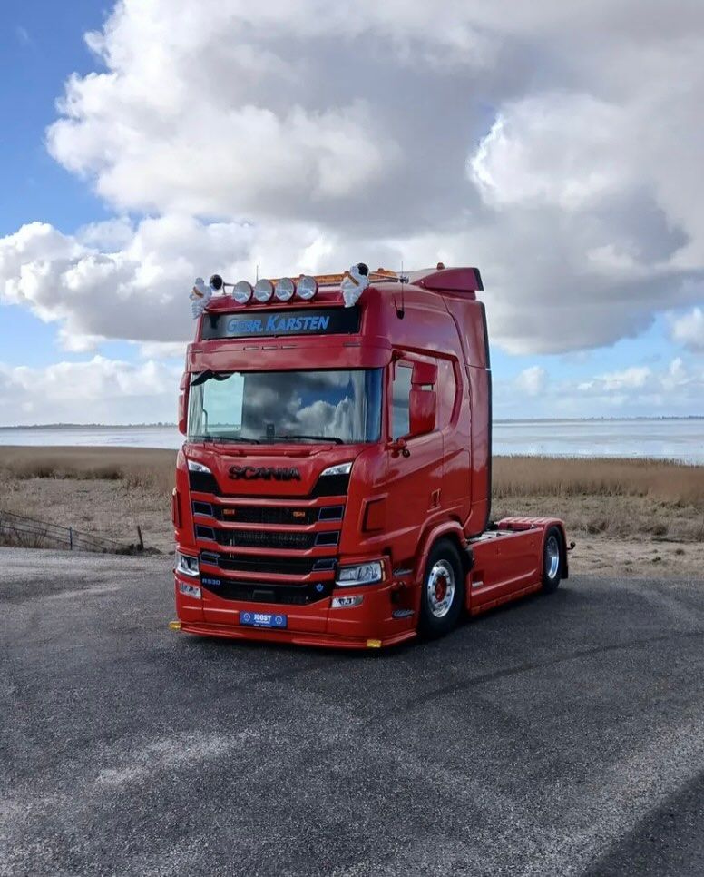 Rode Scania vrachtwagen geparkeerd op een weg vlakbij het water, onder een bewolkte blauwe lucht.