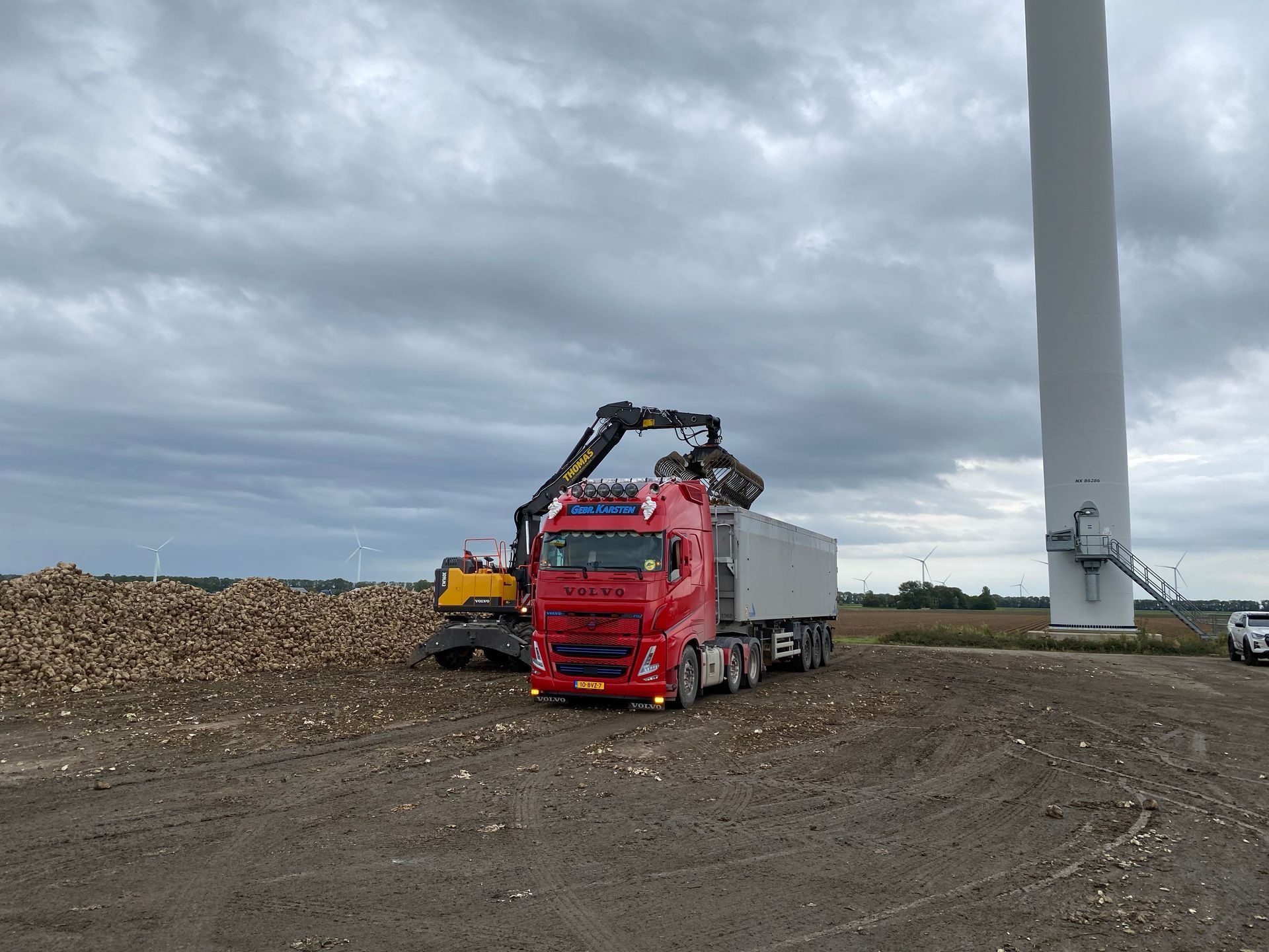 Een rode vrachtwagen wordt door een gele graafmachine geladen met materiaal bij een windturbine op een bewolkte dag.