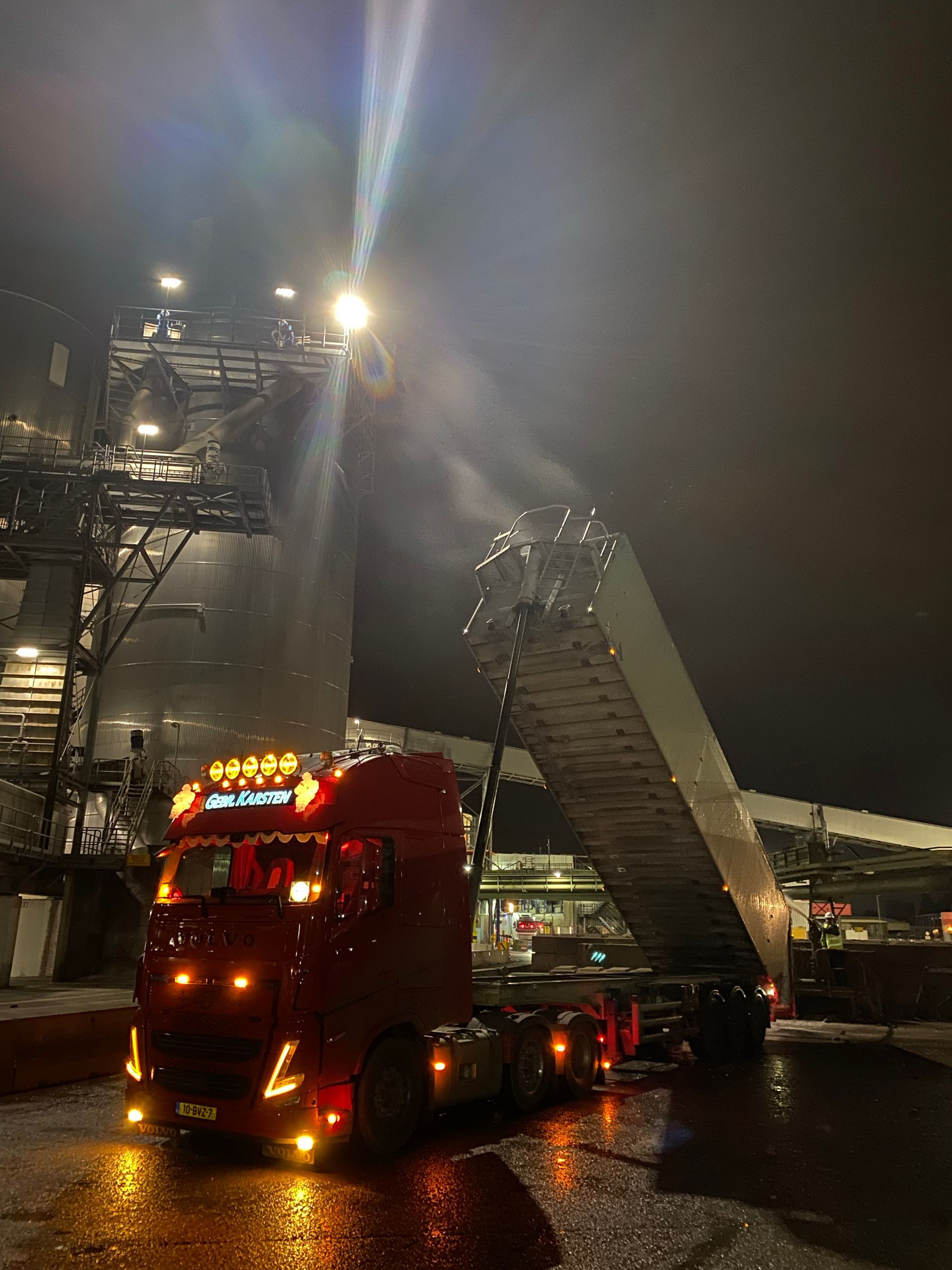 Rode vrachtwagen onder felle verlichting in de nacht, die materiaal lost op een verhoogde constructie.