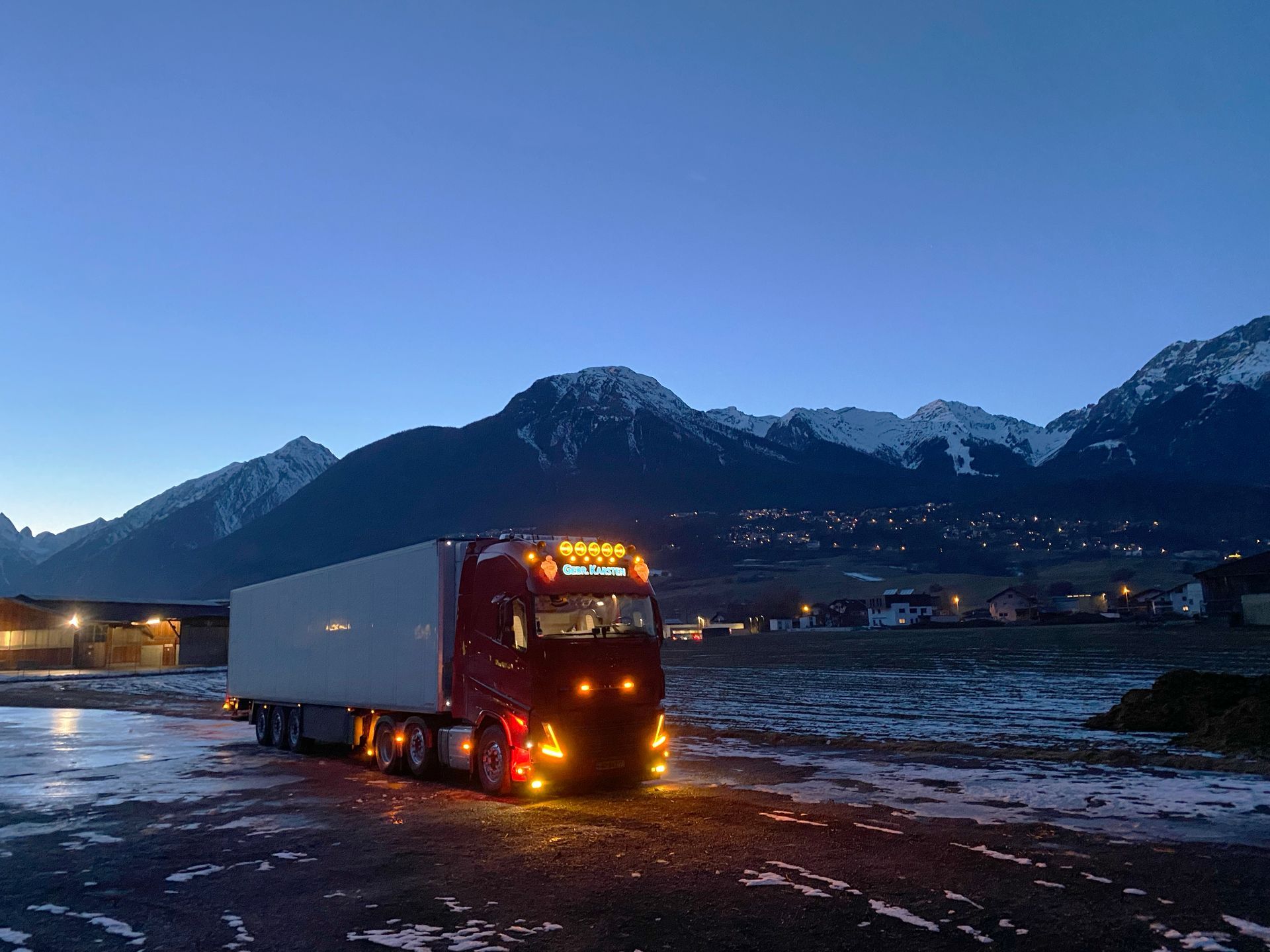 Rode vrachtwagen met aanhanger geparkeerd in de schemering, bergen op de achtergrond, verlichte lichten.
