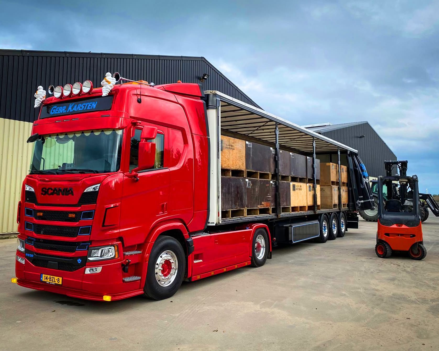 Een rode Scania-vrachtwagen vol dozen, naast een vorkheftruck, tegen een achtergrond van een gebouw en een bewolkte lucht.