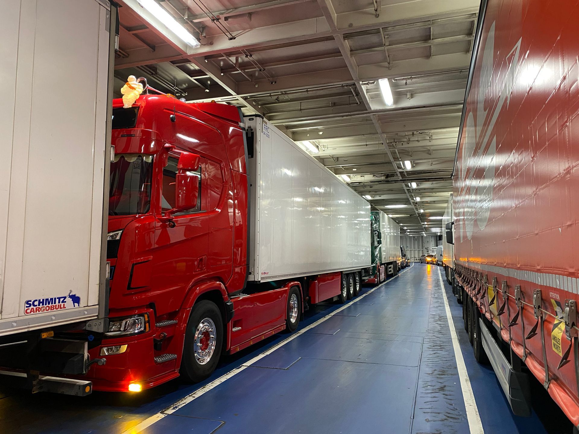 Een rode vrachtwagen geparkeerd in een veerboot, naast andere vrachtwagens in een vrachtruim. TL-lampen schijnen erboven.