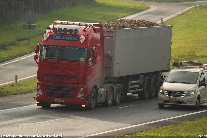 Rode Volvo-vrachtwagen met vracht op de weg, gevolgd door een witte bestelwagen.
