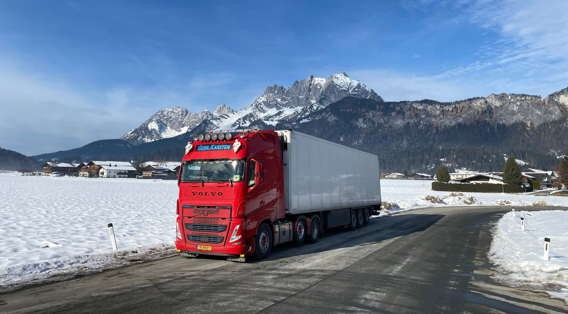 Rode vrachtwagen rijdt op een weg in een besneeuwd landschap met bergen op de achtergrond.