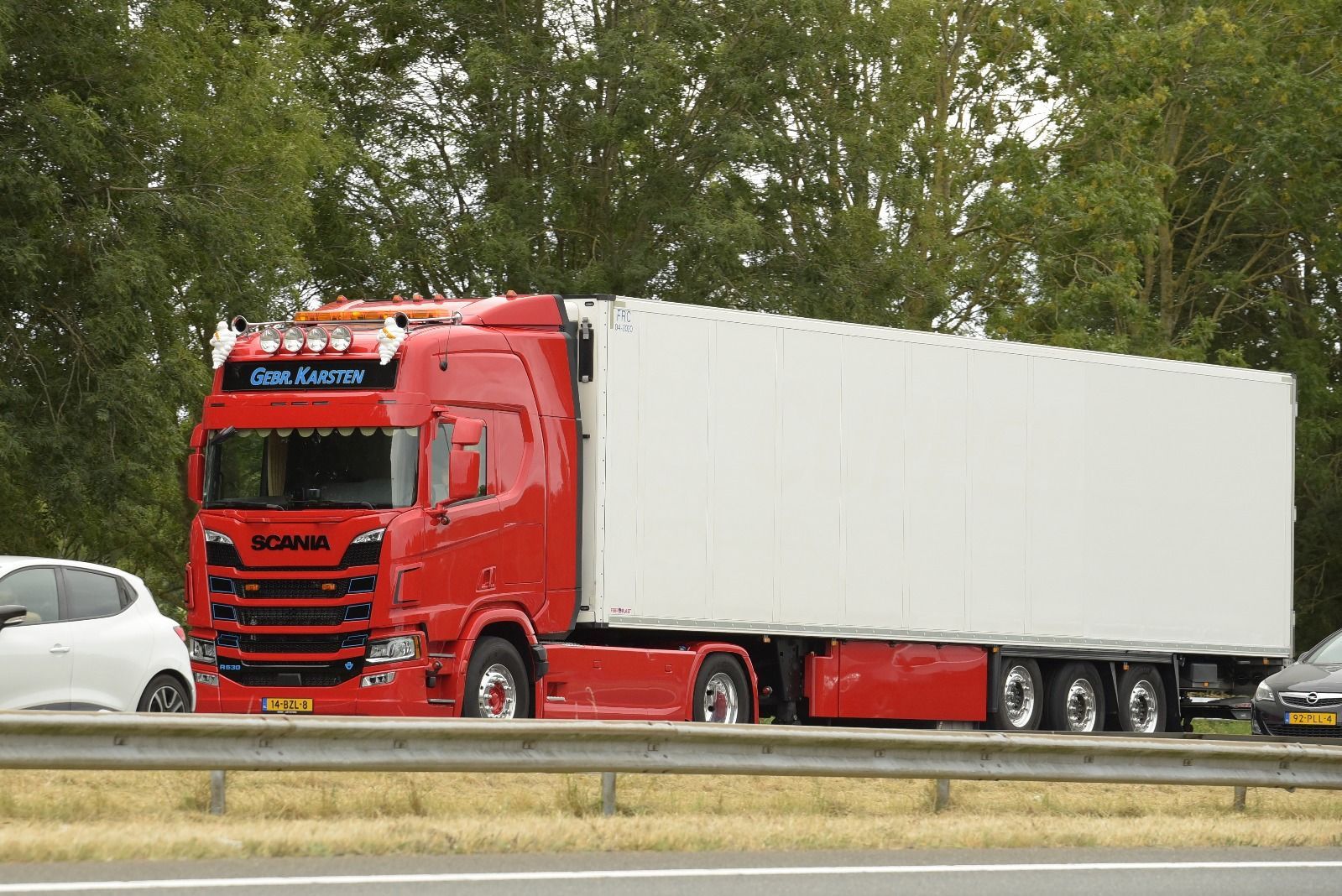 Rode Scania vrachtwagen met witte oplegger op de snelweg; bomen op de achtergrond.