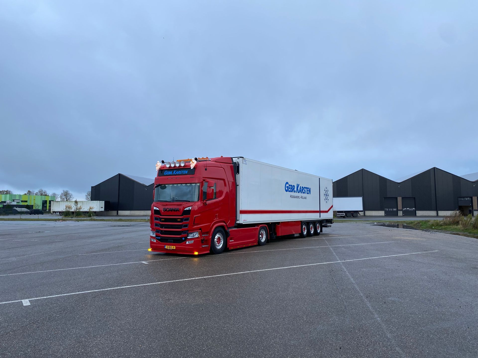 Een rood-witte vrachtwagen op een parkeerplaats op een bewolkte dag. Gebouwen op de achtergrond.