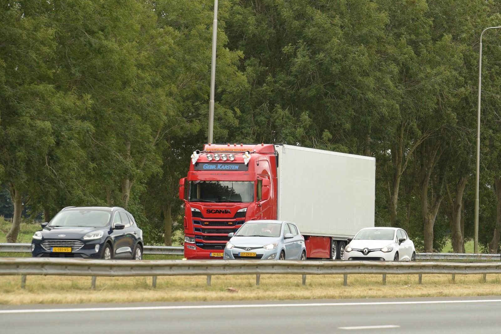 Rode vrachtwagen met witte aanhanger op de snelweg, drie auto's voorbijrijdend. Bomen op de achtergrond.