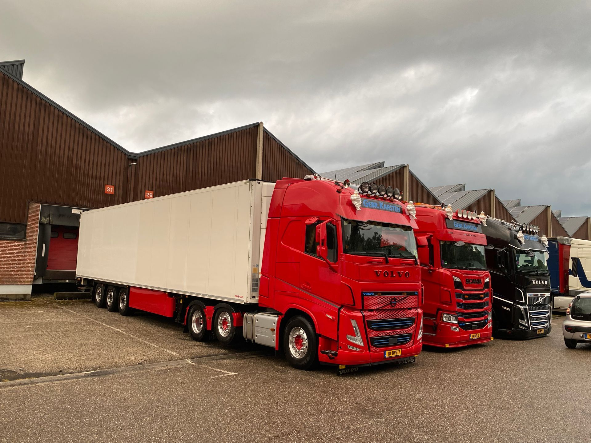 Rode vrachtwagen met aanhanger geparkeerd naast twee andere vrachtwagens. Gebouw op de achtergrond. Bewolkte lucht.