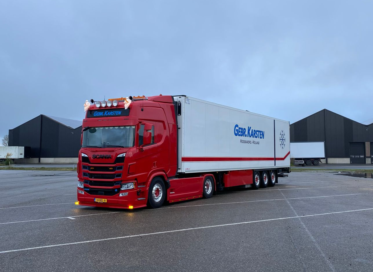 Rood-witte vrachtwagen met oplegger geparkeerd op een grijze ondergrond. De oplegger is bedrukt met de naam